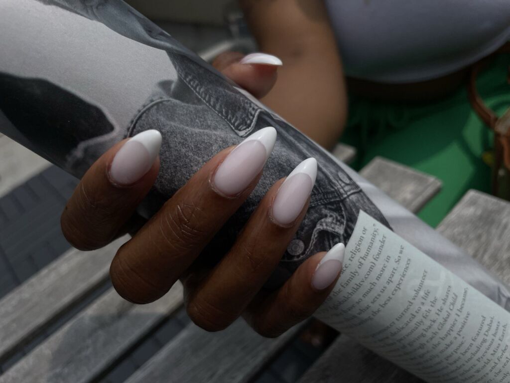 A hand with long, almond-shaped milky white gel nails holding a rolled magazine outdoors.