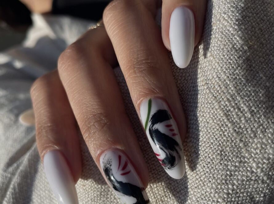 A hand with long almond-shaped white gel nails, featuring abstract black and red brushstroke nail art on two fingers.
