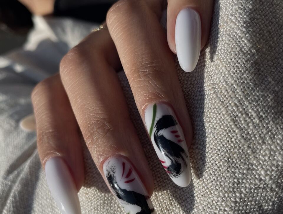 A hand with long almond-shaped white gel nails, featuring abstract black and red brushstroke nail art on two fingers.