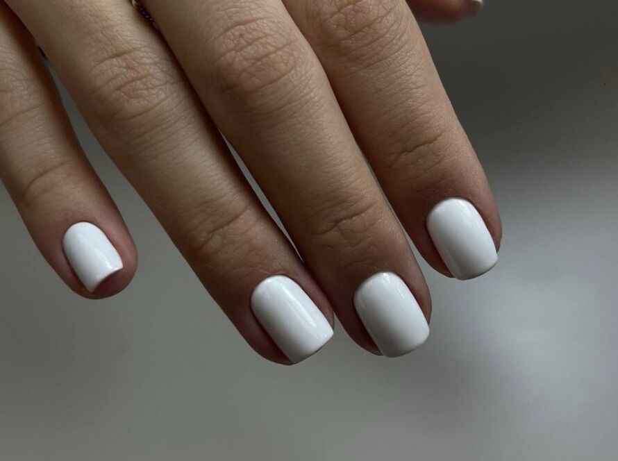 Hand with twisted silver ring displaying short rounded nails with Russian manicure and matte white gel polish against neutral background