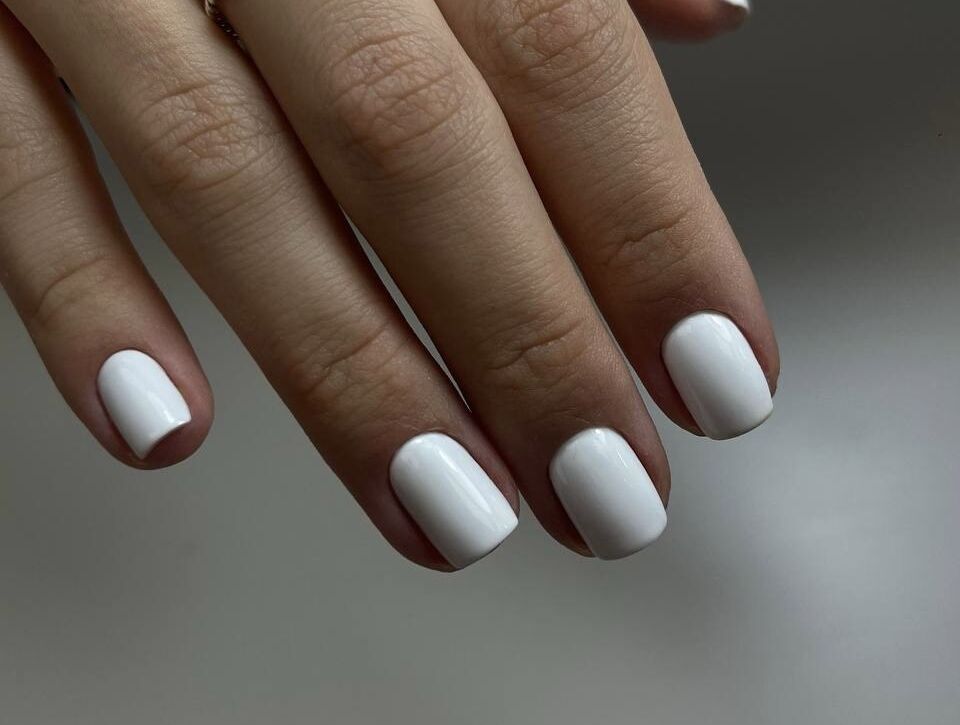Hand with twisted silver ring displaying short rounded nails with Russian manicure and matte white gel polish against neutral background