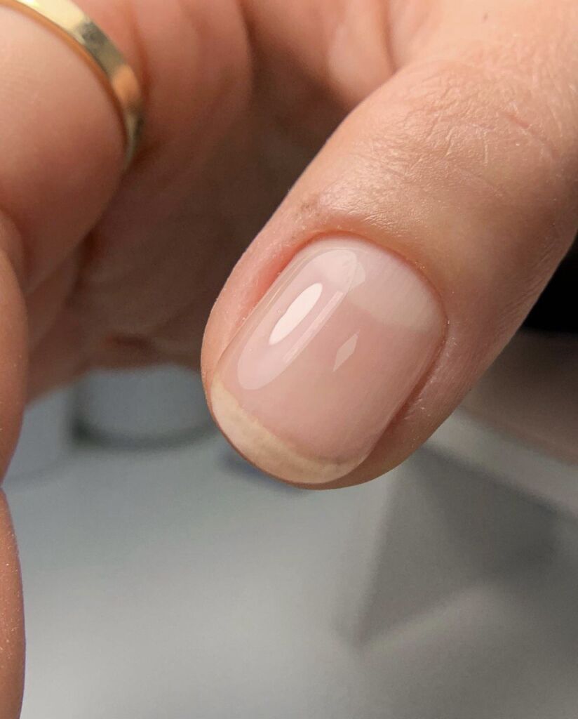 A close-up of a natural short nail with a sheer nude gel polish and a glossy finish.