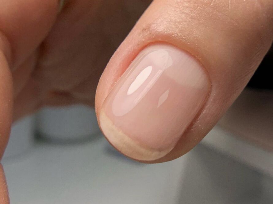 A close-up of a natural short nail with a sheer nude gel polish and a glossy finish.