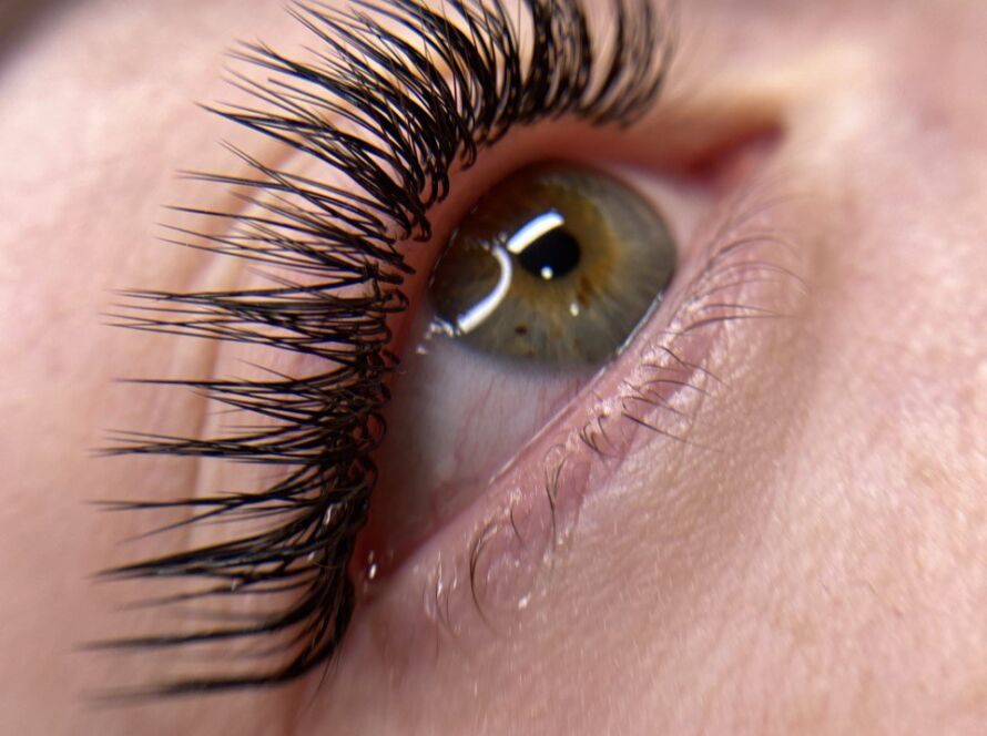 Extreme macro close-up of an open green-hazel eye with a light reflection on the iris, featuring long wispy lash extensions with visible individual strand separation on the upper lash line and sparse natural lower lashes, set against fair, bare skin.