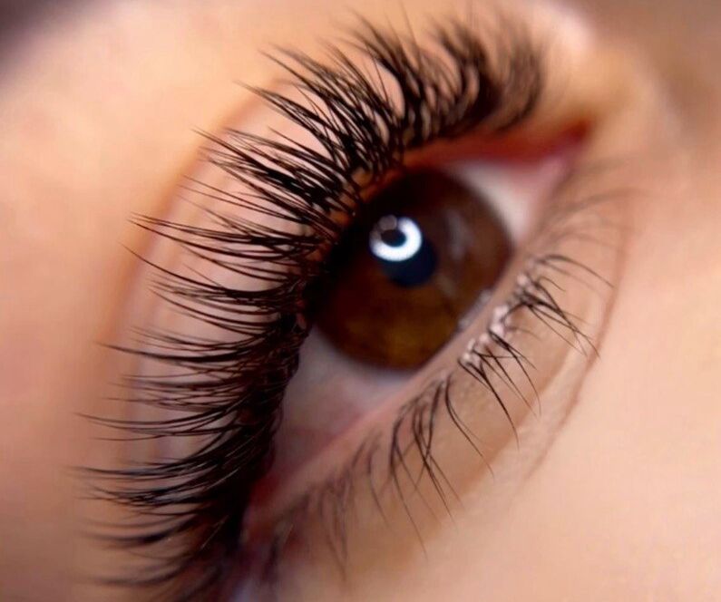 Extreme macro close-up of a brown eye with a light reflection in the pupil, featuring long and full volume lash extensions with a wispy, fanned-out texture on the upper lash line and fine lower lashes, set against soft peachy skin with a partially visible natural brow.