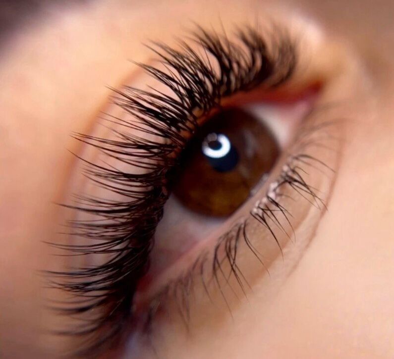 Extreme macro close-up of a brown eye with a light reflection in the pupil, featuring long and full volume lash extensions with a wispy, fanned-out texture on the upper lash line and fine lower lashes, set against soft peachy skin with a partially visible natural brow.