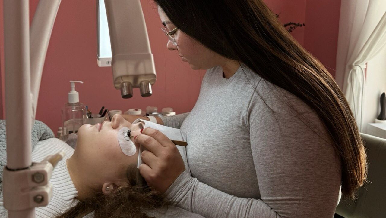 Lash artist with long dark hair applying eyelash extensions to client lying on treatment bed under magnifying lamp in pink salon room