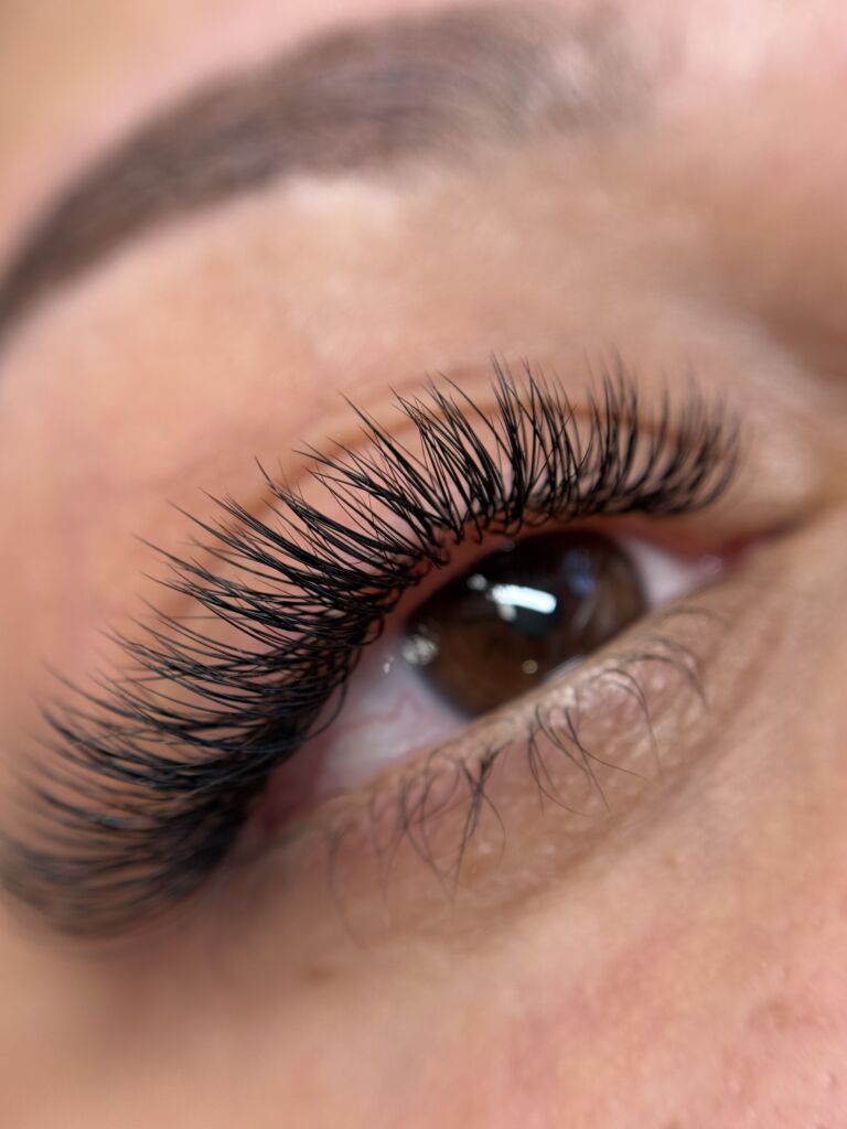 Macro close-up of a brown eye with a light reflection in the pupil, featuring long, full volume lash extensions with a wispy and fluffy texture on both upper and lower lash lines, set against soft peachy skin and a partially visible natural brow.