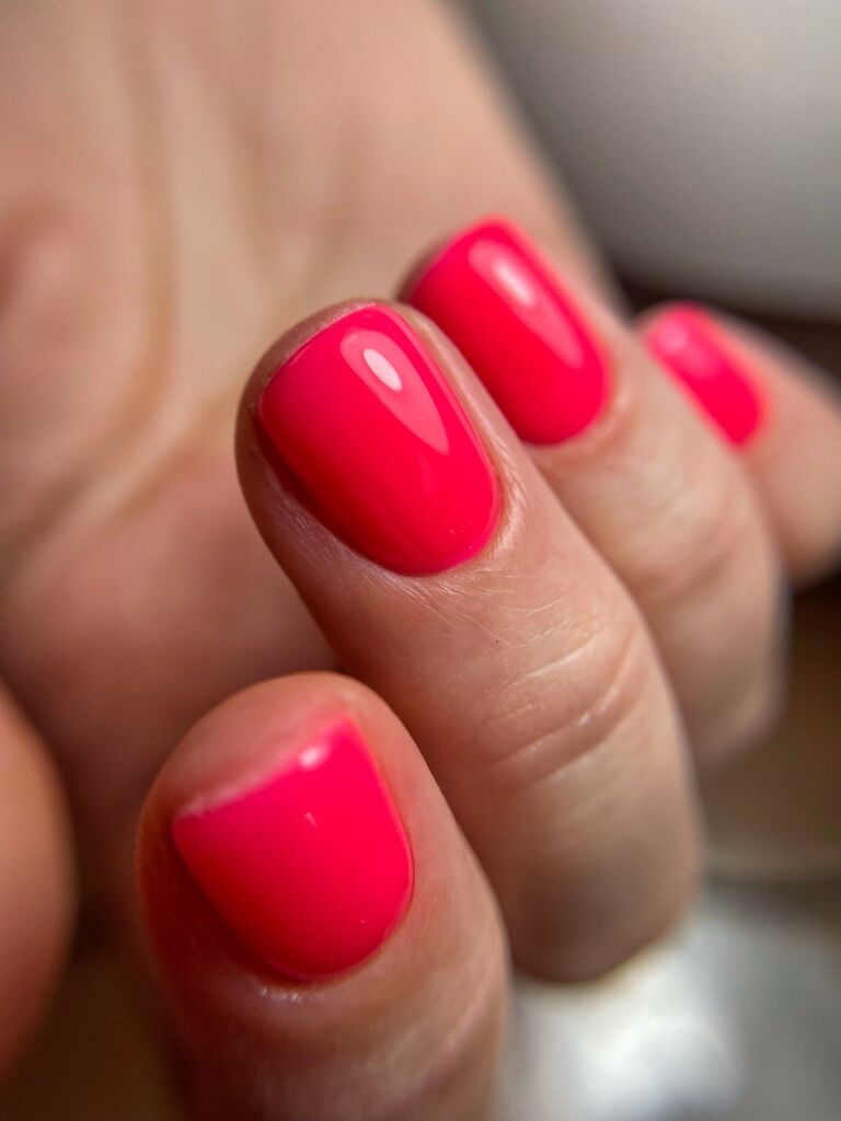 Close-up of short rounded nails with glossy hot coral-pink gel polish manicure showing clean cuticle work and high shine finish