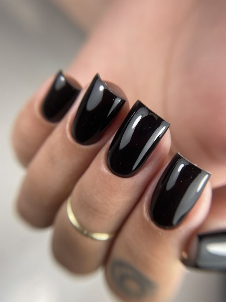 Hand with gold ring displaying medium square-shaped nails with Russian manicure and glossy black gel polish against soft background
