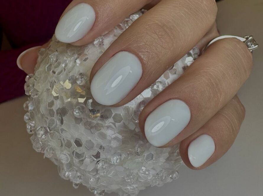 Hand holding crystal decorative ball displaying short oval nails with milky white glossy gel polish and silver rings
