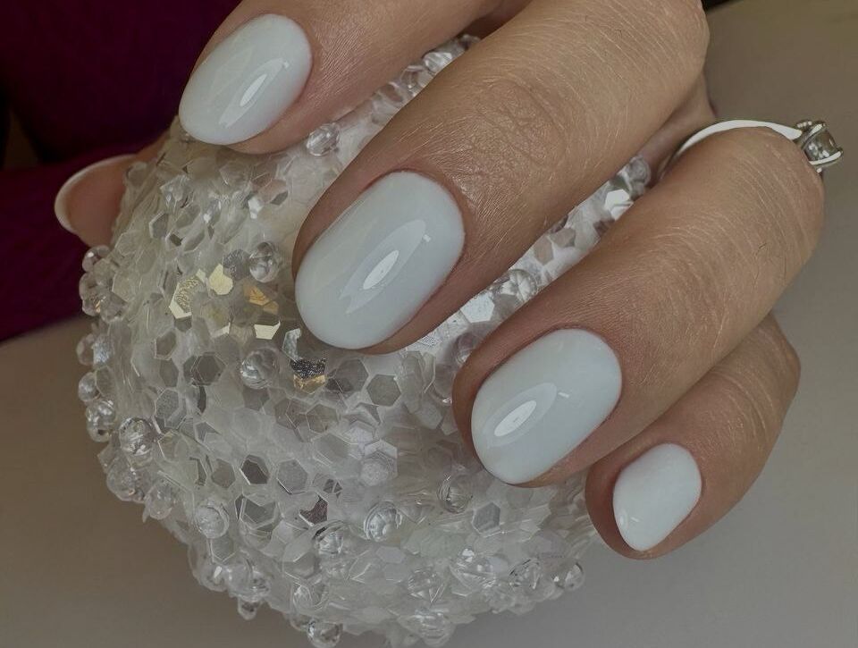 Hand holding crystal decorative ball displaying short oval nails with milky white glossy gel polish and silver rings