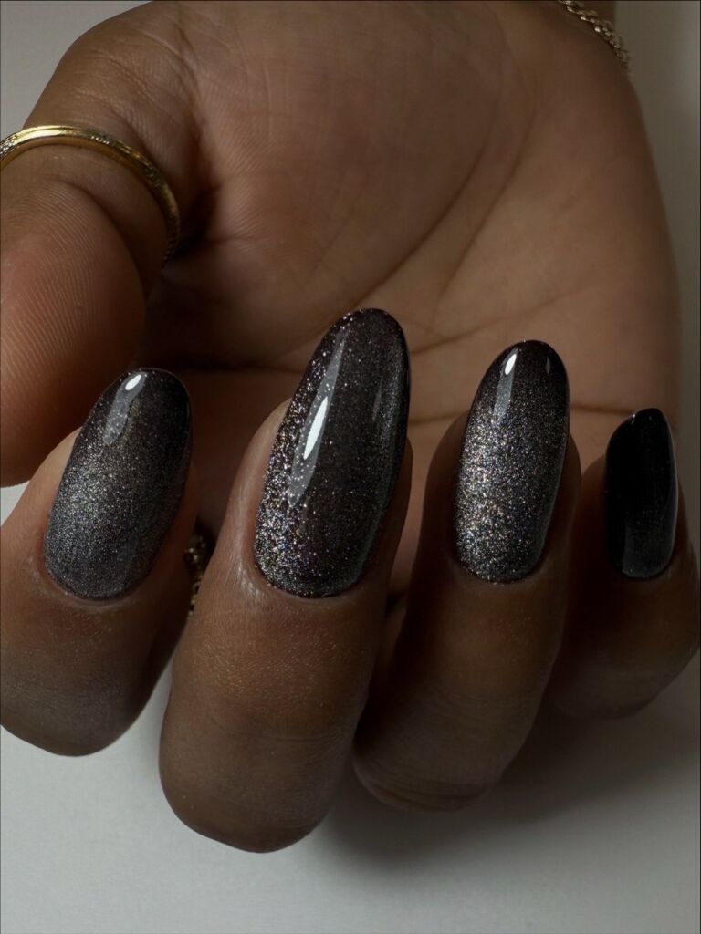 Hand with gold rings displaying long almond-shaped nails with Russian manicure featuring dark charcoal black base with silver glitter shimmer against neutral background