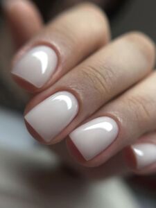 Close-up of a hand with short rounded nails coated in a sheer milky-white high-gloss polish, styled as a clean and delicate bridal manicure against a soft blurred background.