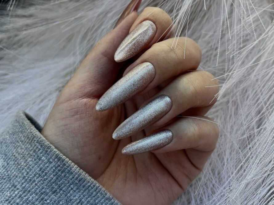 Close-up of long almond-shaped nails with a shimmering champagne silver finish against a soft white textured background.