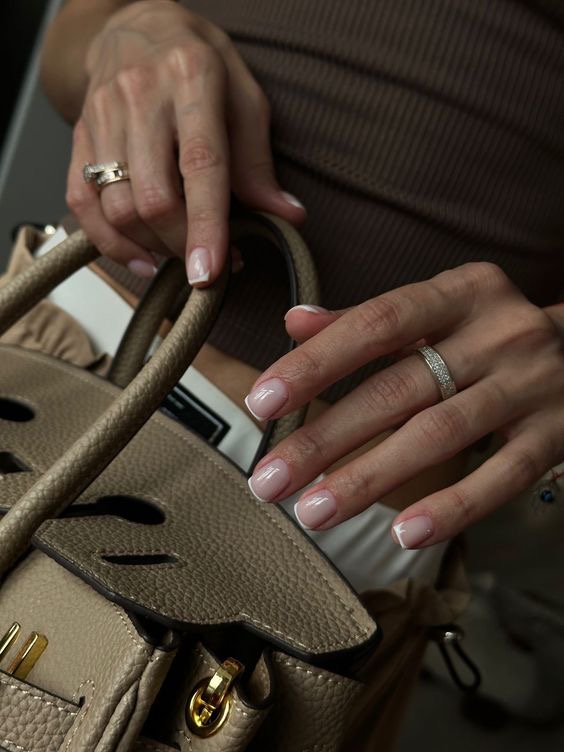 Close-up of short nude French manicure on natural nails near a beige designer-style handbag.