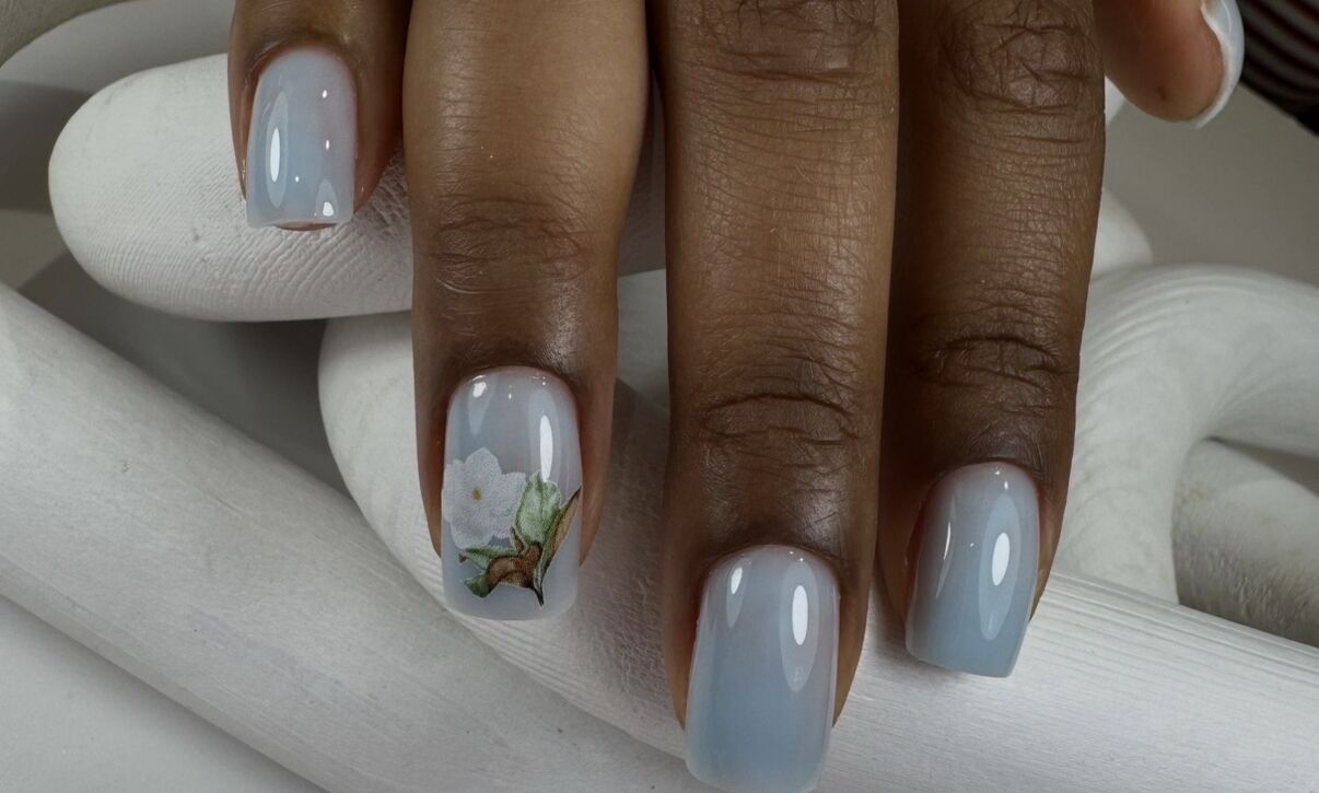 A hand resting on a white sculptural surface displaying short square nails in a soft powder blue glossy gel, with one accent nail featuring a detailed white magnolia flower and green leaf nail art - a refined floral bridal nail design at Midlton Nails Studio in Queens, NYC.