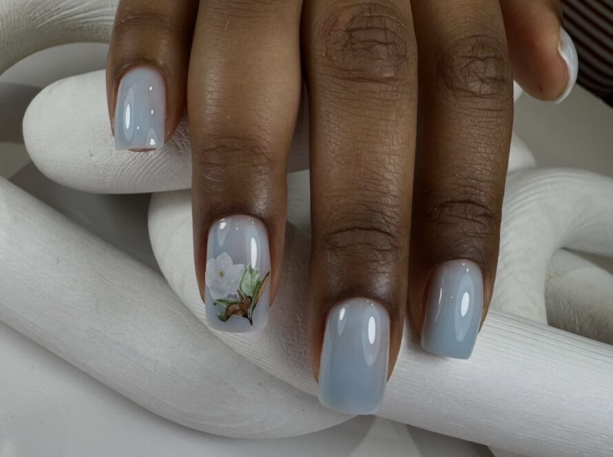 A hand resting on a white sculptural surface displaying short square nails in a soft powder blue glossy gel, with one accent nail featuring a detailed white magnolia flower and green leaf nail art - a refined floral bridal nail design at Midlton Nails Studio in Queens, NYC.