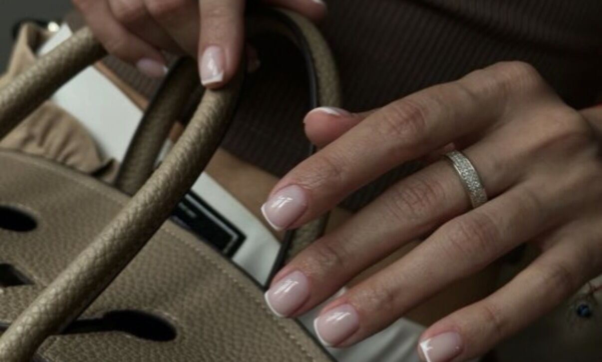 Hands with silver rings holding designer handbag displaying short square-shaped nails with Russian manicure featuring classic French tips with sheer pink base and white tips