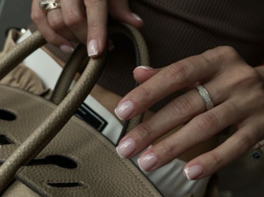 Hands with silver rings holding designer handbag displaying short square-shaped nails with Russian manicure featuring classic French tips with sheer pink base and white tips