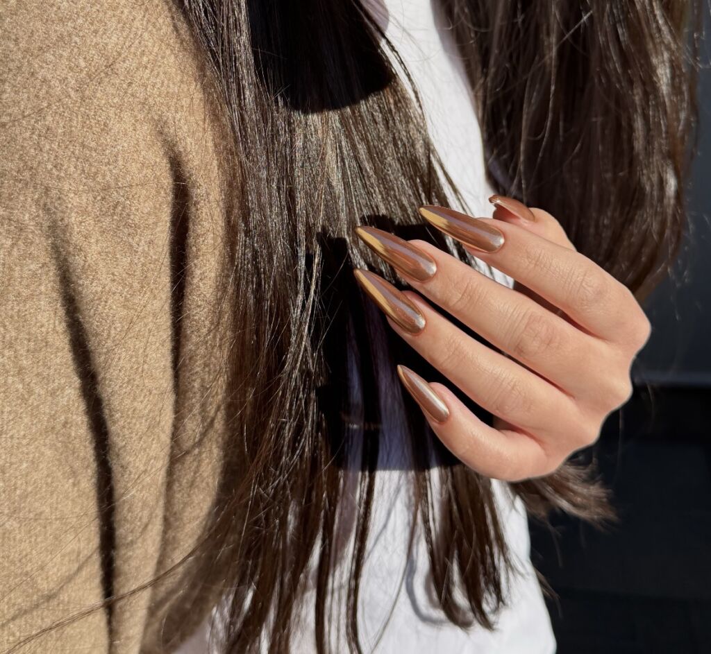 Hand in hair displaying long almond-shaped nails with Russian manicure featuring nude base and metallic gold chrome French tips in natural sunlight