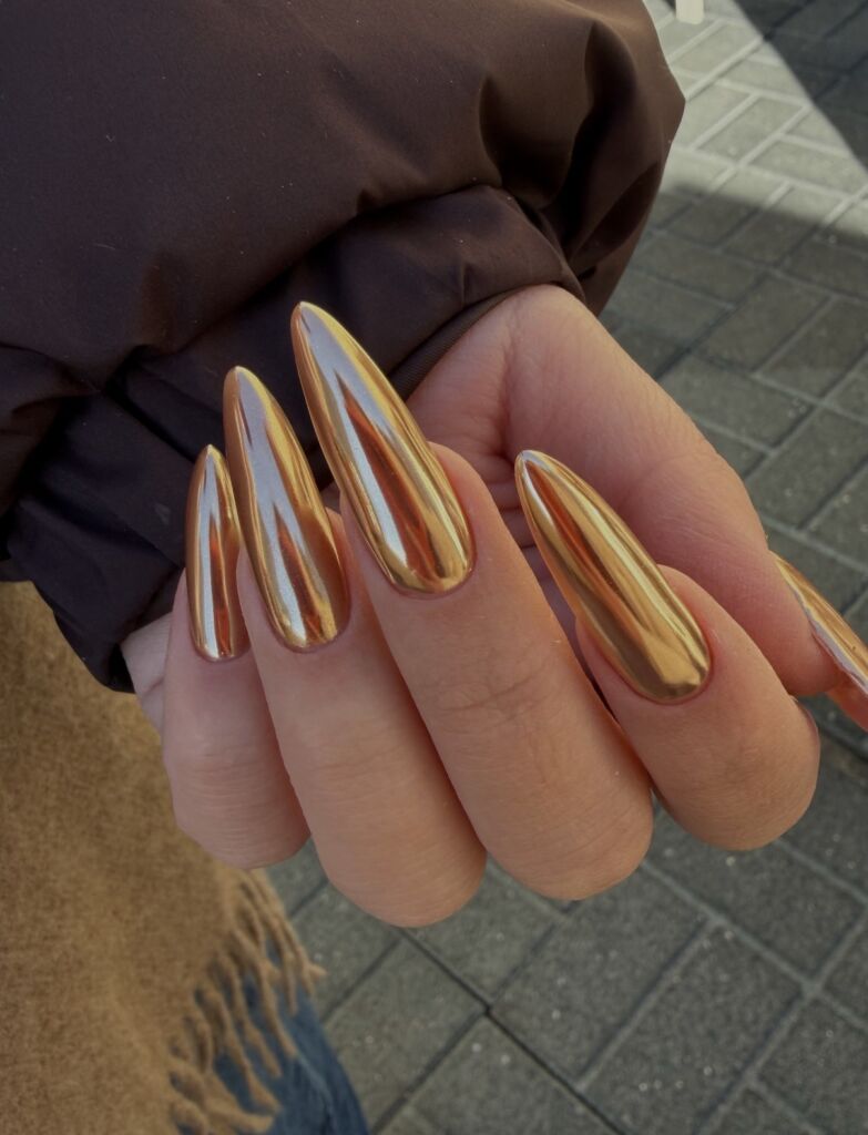 Hand displaying long stiletto-shaped nails with Russian manicure featuring full metallic gold chrome finish with mirror reflection against outdoor setting