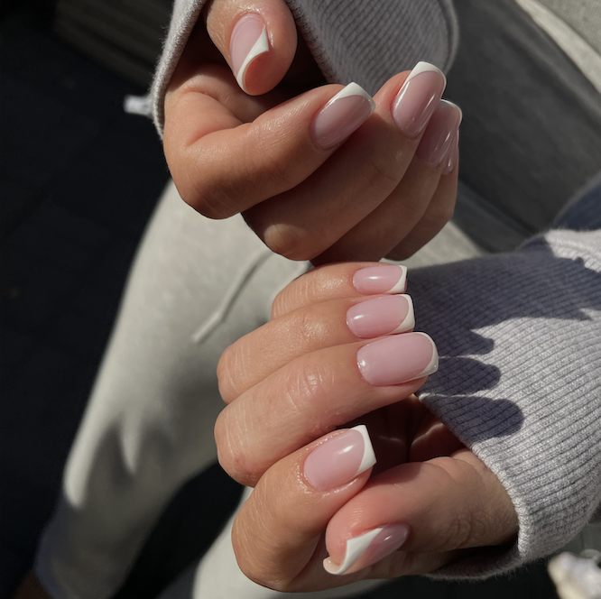 Two hands interlocked outdoors in natural sunlight, wearing a gray ribbed sweater, displaying short square nails with a sheer pink base and a modern white French tip with a curved swirl detail - a clean and polished contemporary manicure.