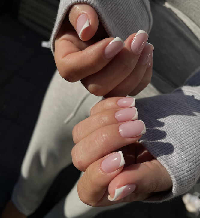 Two hands interlocked outdoors in natural sunlight, wearing a gray ribbed sweater, displaying short square nails with a sheer pink base and a modern white French tip with a curved swirl detail - a clean and polished contemporary manicure.
